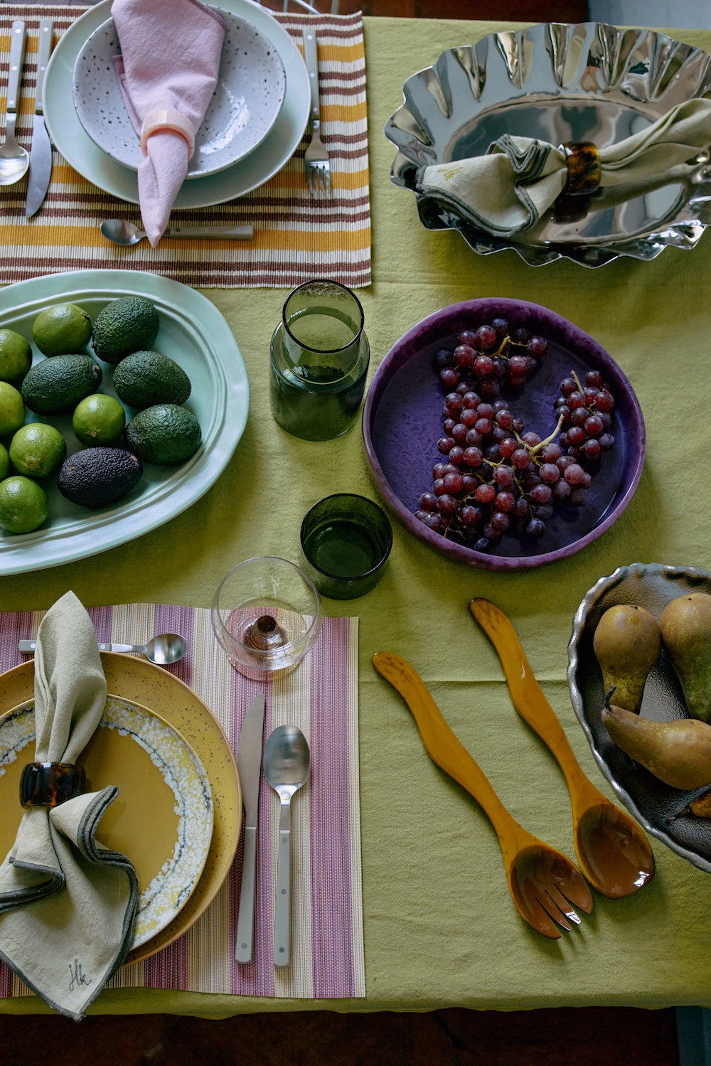 The HKLIVING ochre salad cutlery styled as part of a full table setting on a green linen tablecloth, with grapes, avocados, limes and patterned place settings