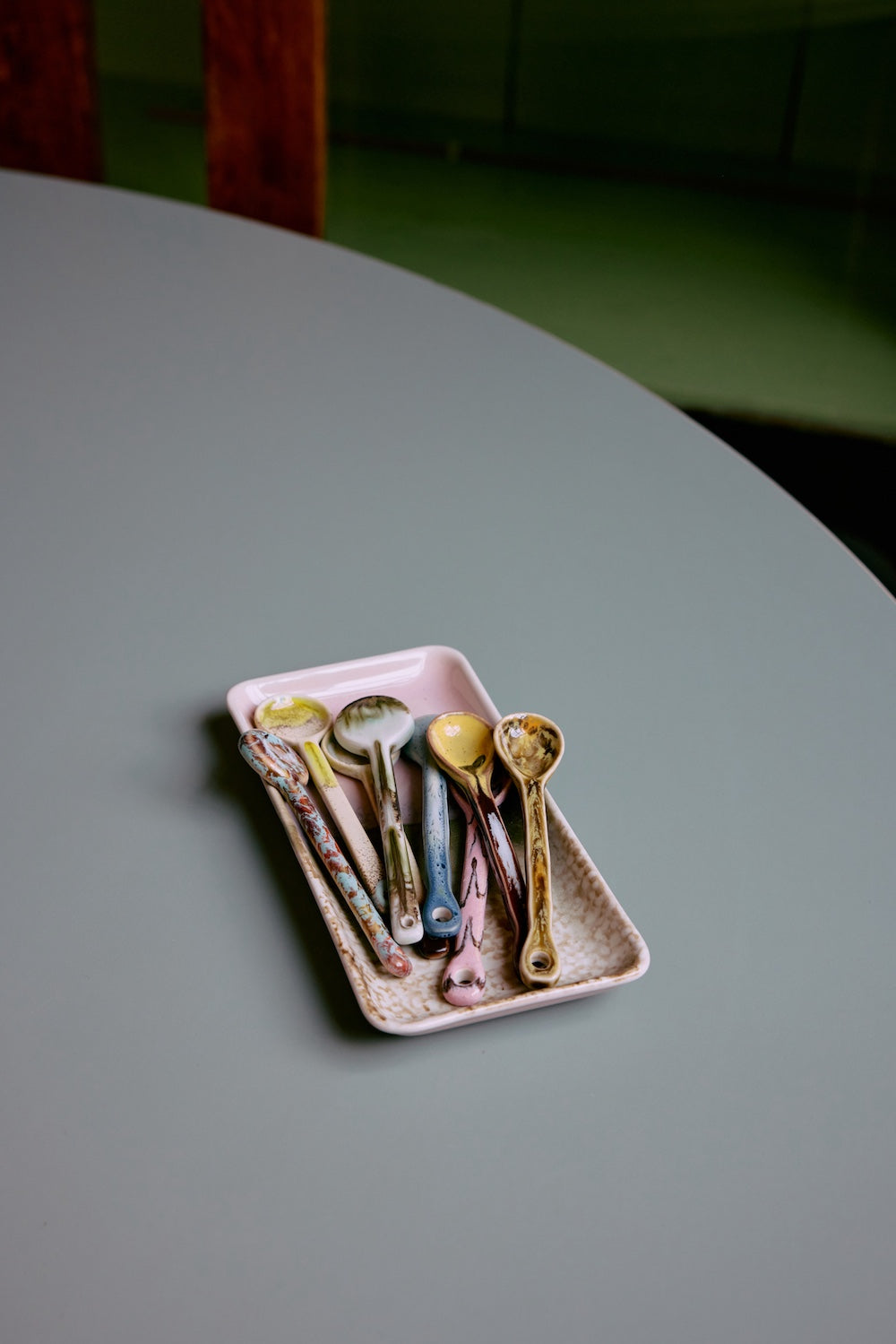 The HKLIVING Sketch spoons, set of 4, arranged on a pink stoneware tray on a round grey table, with a green wall in the background