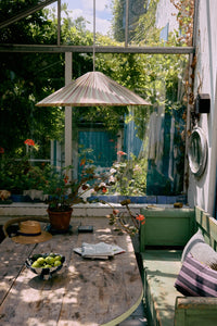 Plisse pendant lamp in Verdura by HKLIVING hanging above a wooden table in a sunlit garden room surrounded by greenery