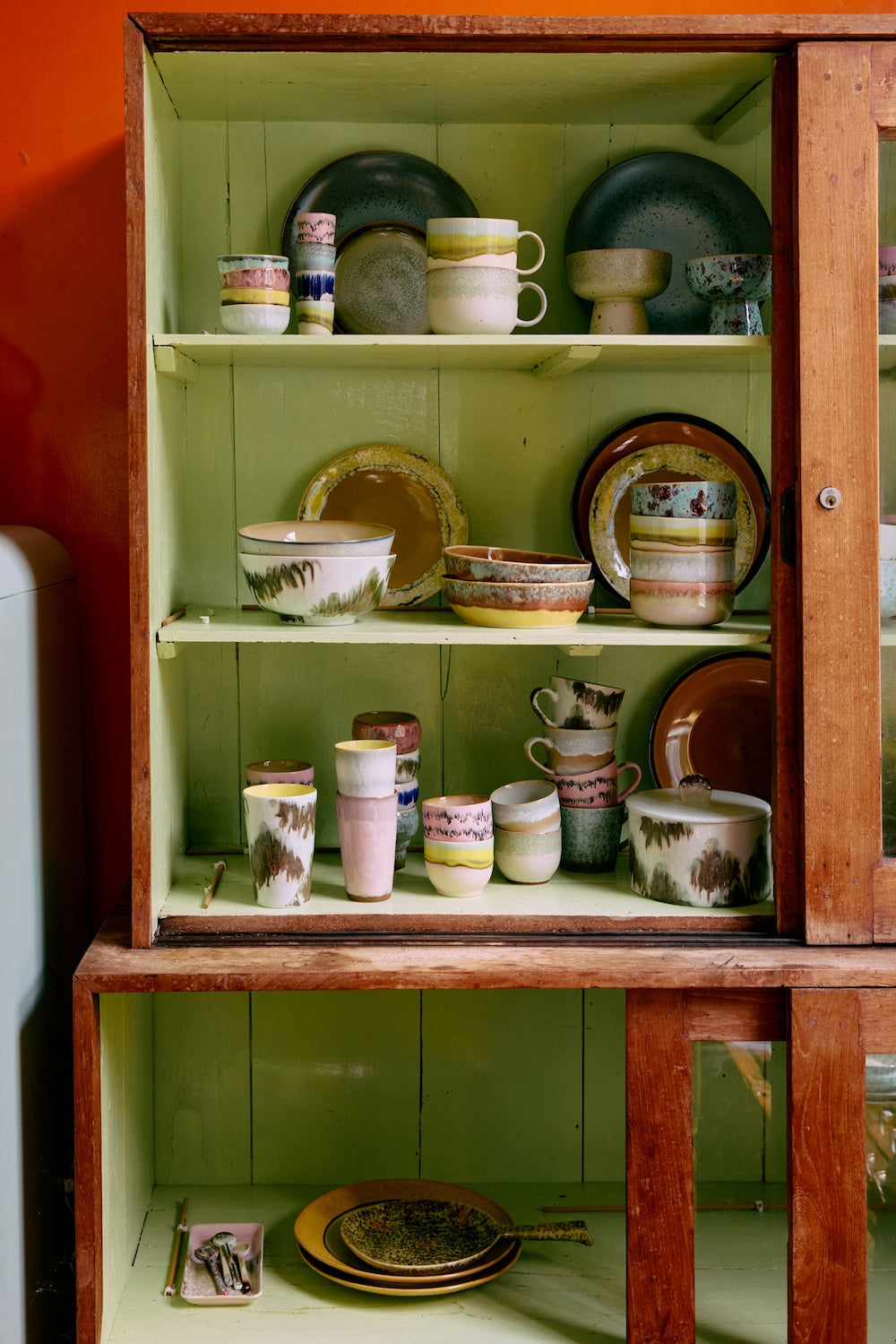 HKLIVING 70s ceramics collection arranged inside a vintage wooden cabinet with sage green shelving, showing an assortment of mugs, bowls and plates