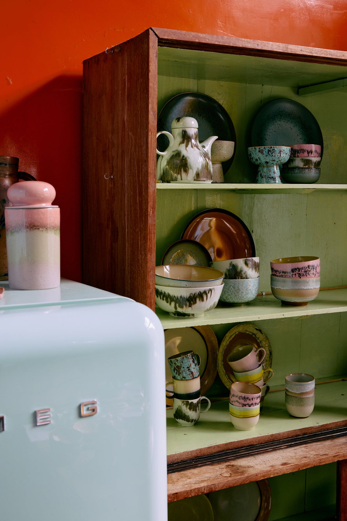 Vintage ceramic items on a wooden shelf against a red wall with a light blue refrigerator.