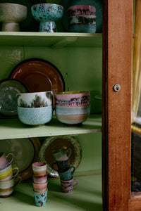 The HKLIVING Futurism ristretto mugs stacked inside a green wooden cabinet, alongside stoneware bowls, plates and jugs from the 70s ceramics collection