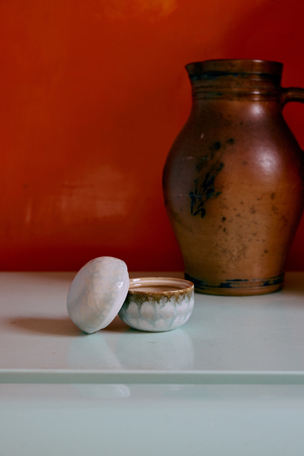 70s Ceramics sugar pot in Petal shown open with its lid resting beside it, styled next to a rustic stoneware pitcher against an orange wall
