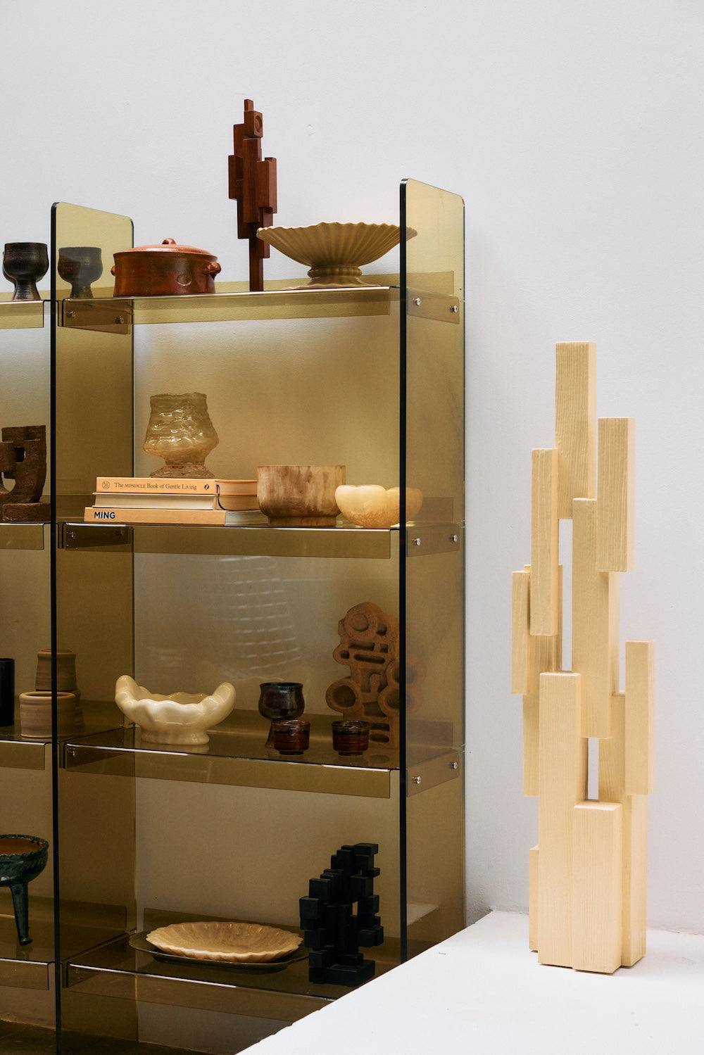 The HKLIVING oyster glass bowl displayed on a glass shelving unit alongside ceramic vessels, sculptural objects and a stack of books