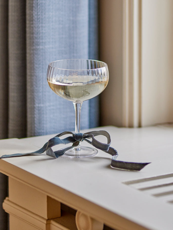 Astrid champagne coupe glass with ribbed design, filled with wine and tied with ribbon, on a windowsill
