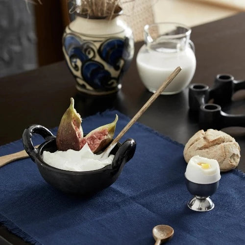 lovely breakfast set up with greek yoghurt and a fresh fig cut in half in a black handled bowl with a long wooden spoon set in side ready to eat. A dippy egg with the top removed and a small roll sat next to it and a jug with milk in the background