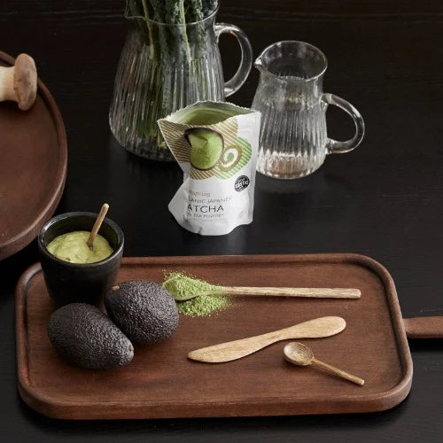 a dark wood tray with 2 avocados (your guess is as good as mine as to why they're there) and a pack of matcha powder. The long spoon has been used to scoop the matcha powder into a mug