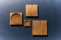 A flat lay of teak pieces from the HKLIVING collection, including the square teak cube alongside two round-carved teak bowls, arranged on a dark blue background