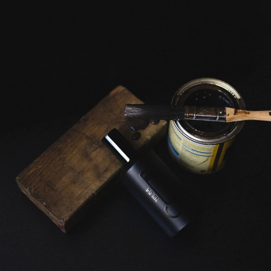 an open can of wood varnish with a brush, recently dipped inside the tin, resting across the top, dripping varnish onto a block of old wood below representing one fo the scents within fischersund number 54