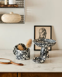 black and white terrazzo bowls and a small dish styled on a white marble counter with a travertine trinket box, a wooden hand mirror and a framed print in the background