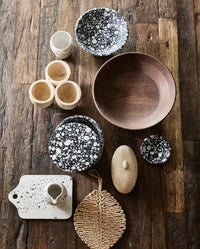 flatlay of black and white terrazzo bowls and dishes by Nordal on a dark wood surface, alongside a walnut wood salad bowl, small stone cups, a travertine trinket box, a speckled ceramic jug on a matching board, and a woven leaf-shaped trivet