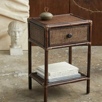 a small green, snady storage bowl sat atop a small, dark brown, wicker and cane drawer unit, with books on a lower shelf, and a marble head in the background on a small pedastal