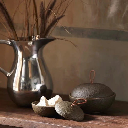 A wooden sideboard with 2 green lidded bowls, both with a leather strap lid hook, one with the lid off revealing sweets inside and a silver jug with dried grasses in the background