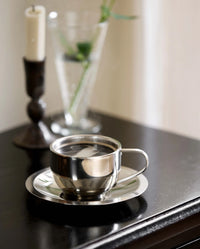 Close-up of the VIREA chrome cup and saucer by Nordal on a dark wood table with a candlestick and vase behind