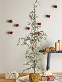 Frosted green Vita tree with a moss base beside wrapped gifts and a dark bench, with red paper ornaments hanging on the wall behind