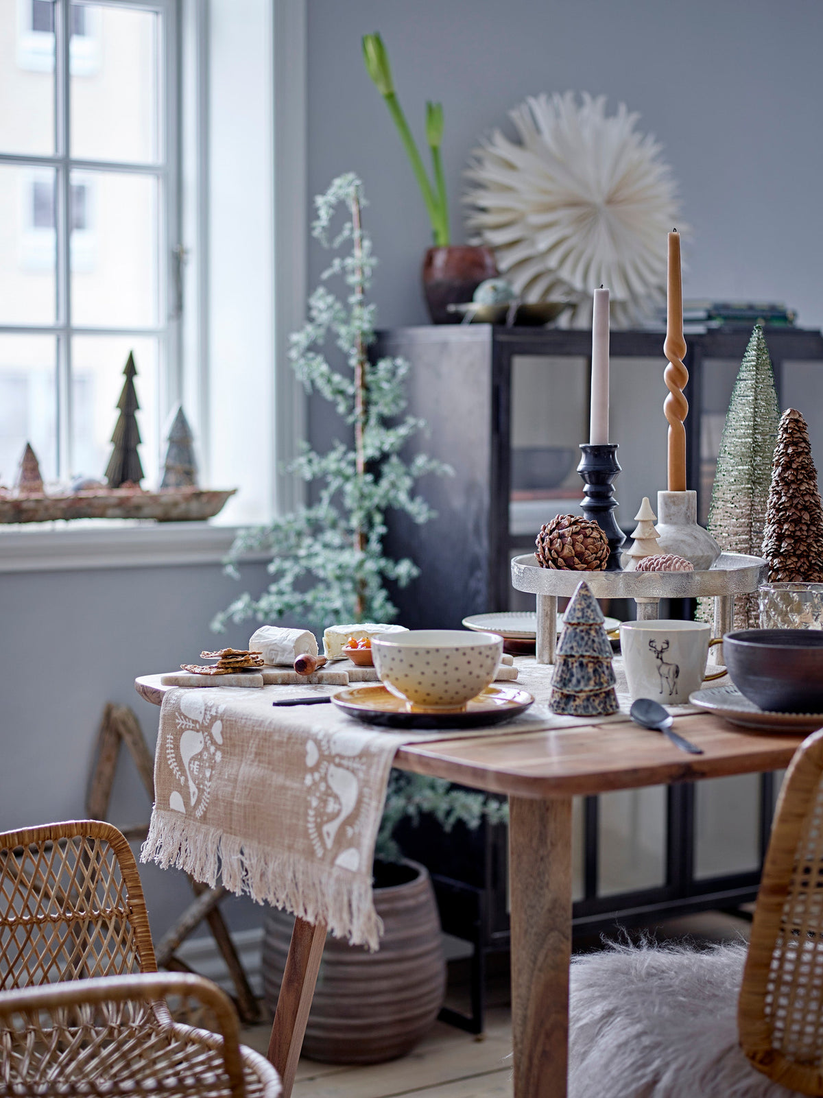 Vita tree standing by a window beside a Christmas dining table set with candles, pine cone decorations and rattan chairs