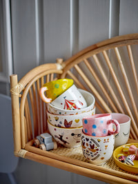 Stacked ceramic cups with colorful designs on a wicker shelf against a gray wall.