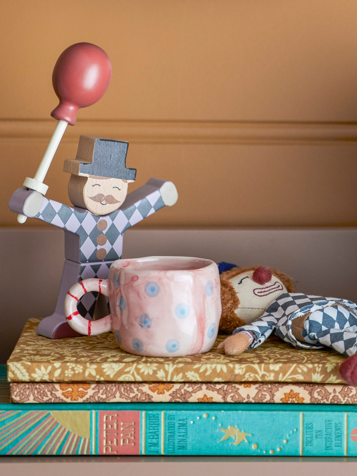 Wooden figurine with a top hat and balloon, sitting on a pink mug with books underneath.