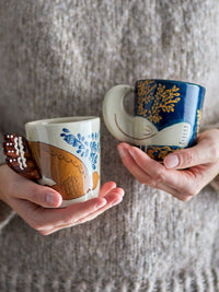 Hands holding two Bloomingville Hezha stoneware cups with raised bird-wing handles in nature and blue colours.