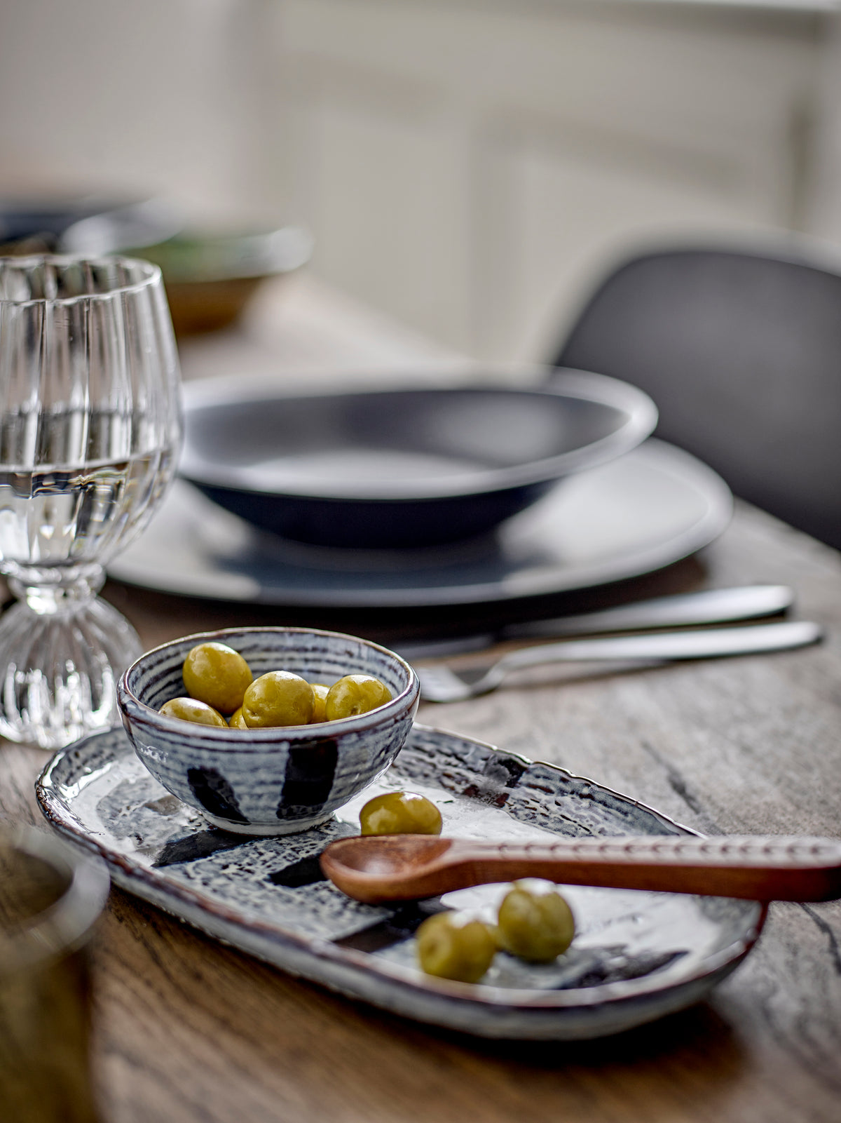 Taurin ribbed wine glass on a wooden table beside dark plates, a bowl of green olives and a patterned tray