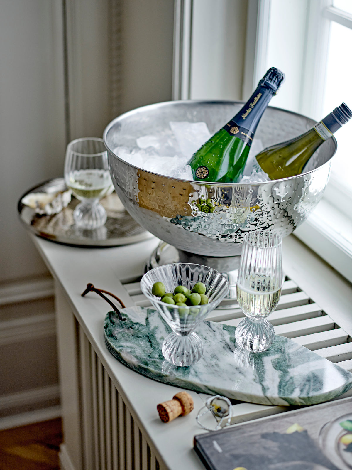 Silver ice bucket with bottles of champagne on a marble tray with glasses and olives.