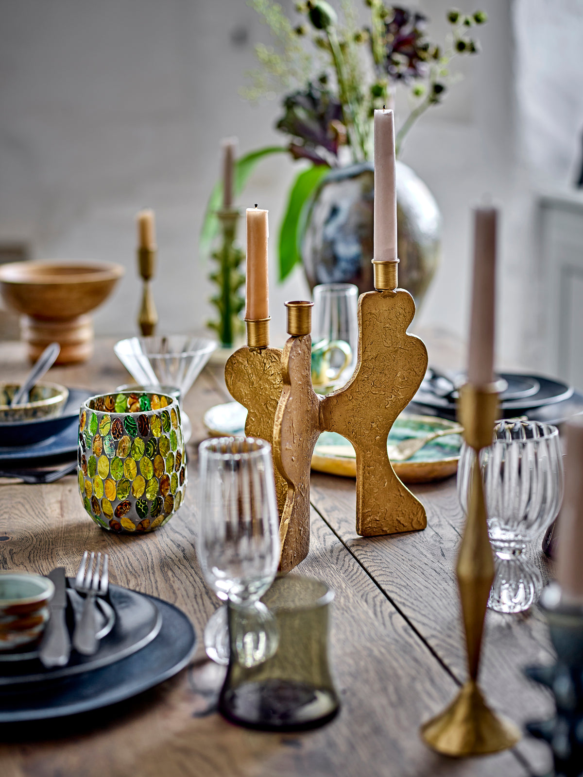 Taurin glassware on a rustic wooden table set with dark plates, brass candlesticks with tapers and a vase of dried stems