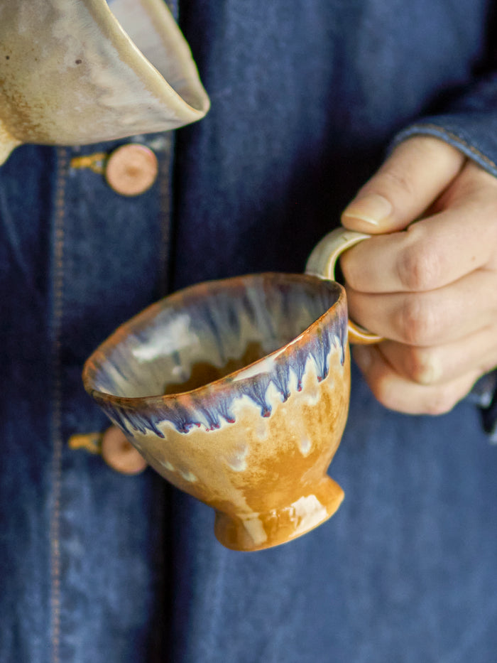 Imogen cup in blue and brown reactive glaze stoneware, handmade, held in hand
