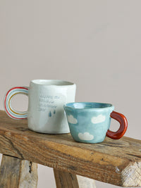 blue nini cup with cloud pattern and red handle next to rainbow mug on rustic bench
