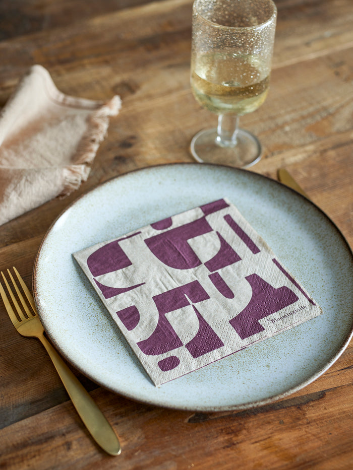 Olica napkin with burgundy graphic print, styled on a plate with gold cutlery, natural wood table
