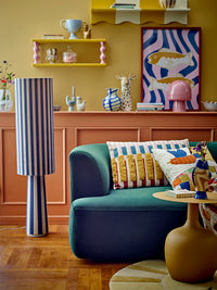 Lane Cushion styled on a green sofa in a colourful room with striped lamp and wall shelves
