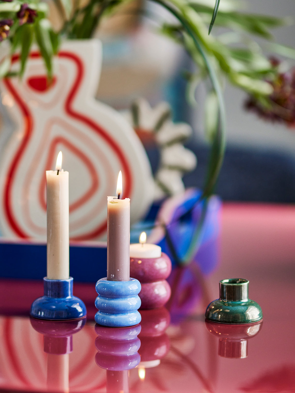 Four Tana candle holders in blue, dusty pink and green holding lit taper candles and a tea light on a glossy pink table
