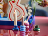 Wide view of the Tana candle holders with lit candles on a glossy pink table in front of a red and white vase