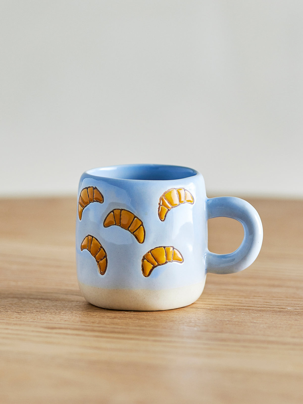 Millie light blue cup with orange hand-painted croissants on a light oak table against a soft grey wall