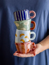 Hand holding a stack of four colourful hand-painted mini cups in front of a blue top, with the croissant cup third from the top