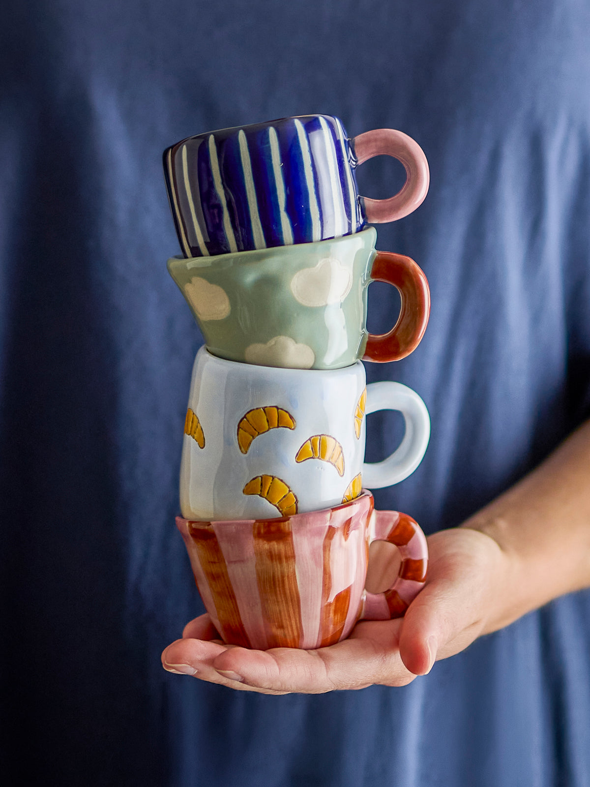 Hand holding a stack of four colourful hand-painted mini cups in front of a blue top, with the croissant cup third from the top