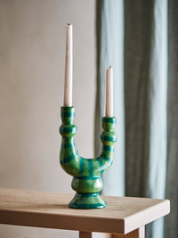 Salandy green striped candle holder with two unlit taper candles on a pale wooden console table in front of a grey curtain