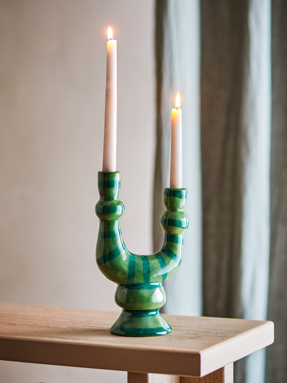 Salandy green striped candle holder with two lit taper candles on a pale wooden console table in front of a grey curtain