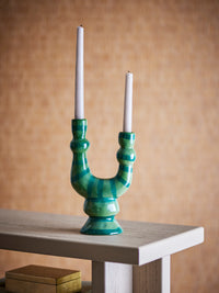 Salandy green and teal striped candle holder with two unlit taper candles on a pale wooden table against a warm ochre wall