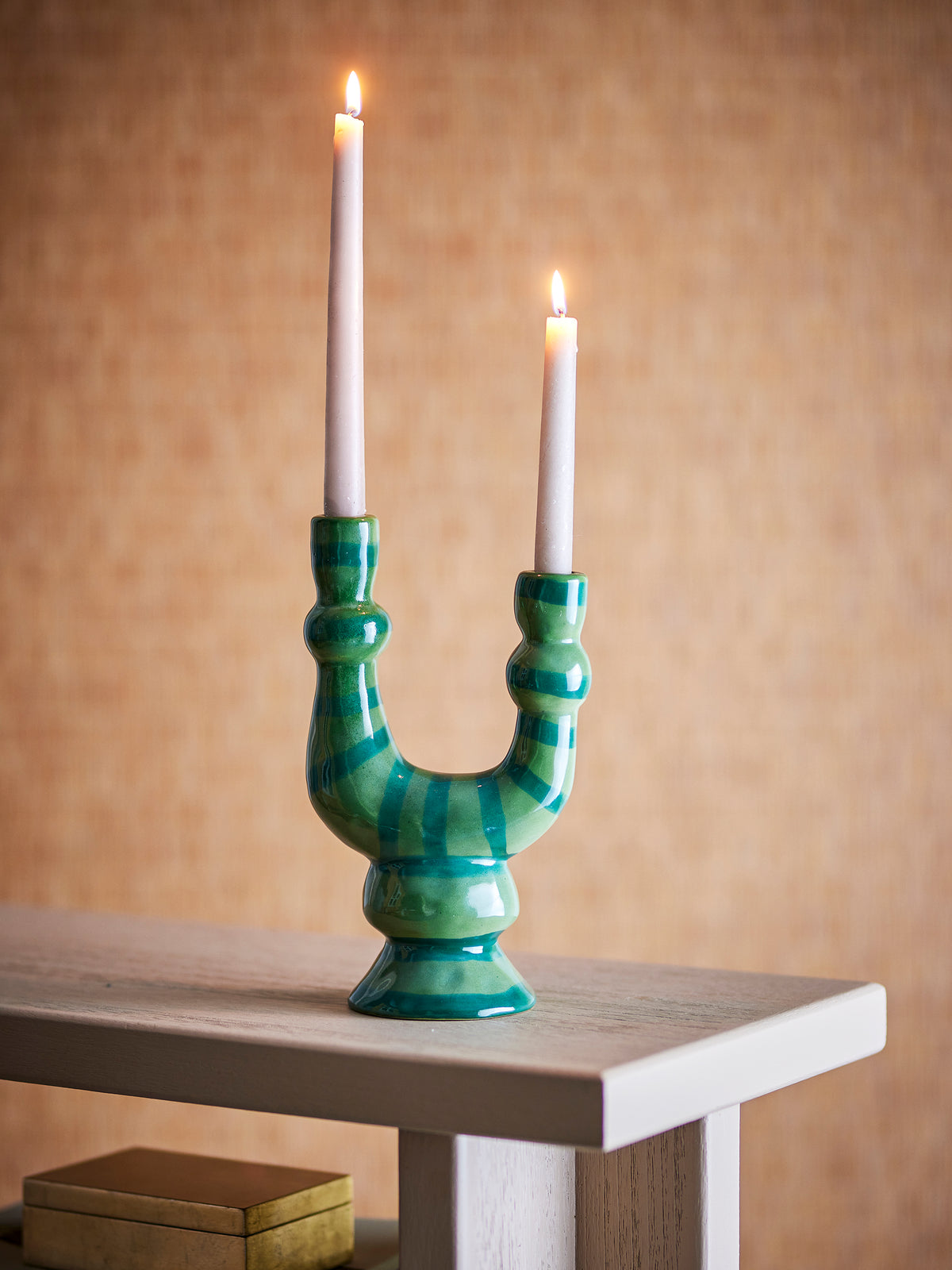 Salandy candle holder with two lit taper candles on a pale wooden table against a warm ochre wall