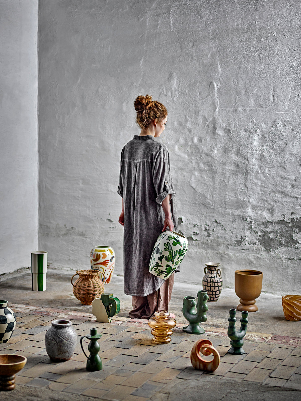 Woman in a grey linen shirt standing among ceramics and vases on a tiled floor, with green sculptural candle holders among the pieces