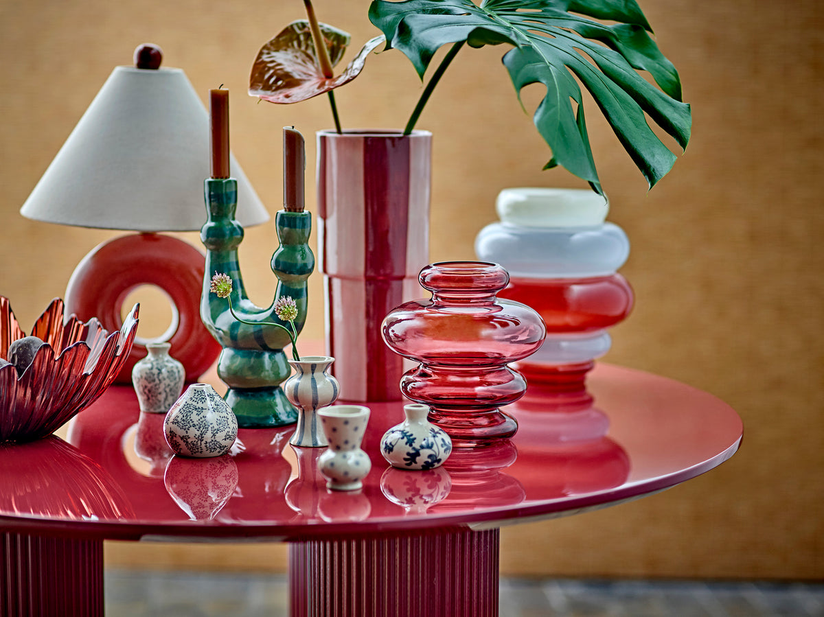 Salandy candle holder with two burgundy taper candles on a round red table beside glass vases, a table lamp and a monstera leaf