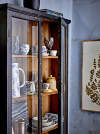 Hazem cabinet styled with blue and white patterned jugs, mugs and bowls, a cut-glass wine goblet and a stoneware jar visible through the open glazed door