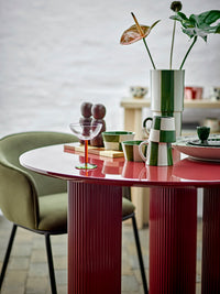 Round red dining table set with green stoneware, a coupe glass and a vase of foliage, with the Salvi mills at the back
