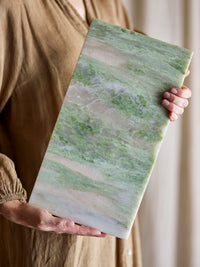 Close-up of the Keika green marble serving board held in both hands, showing the natural stone's grey, green and blush marbling in detail