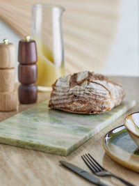Keika green marble serving board styled on a kitchen table with a loaf of sourdough bread, wooden pepper mills, a glass oil cruet and stoneware plates