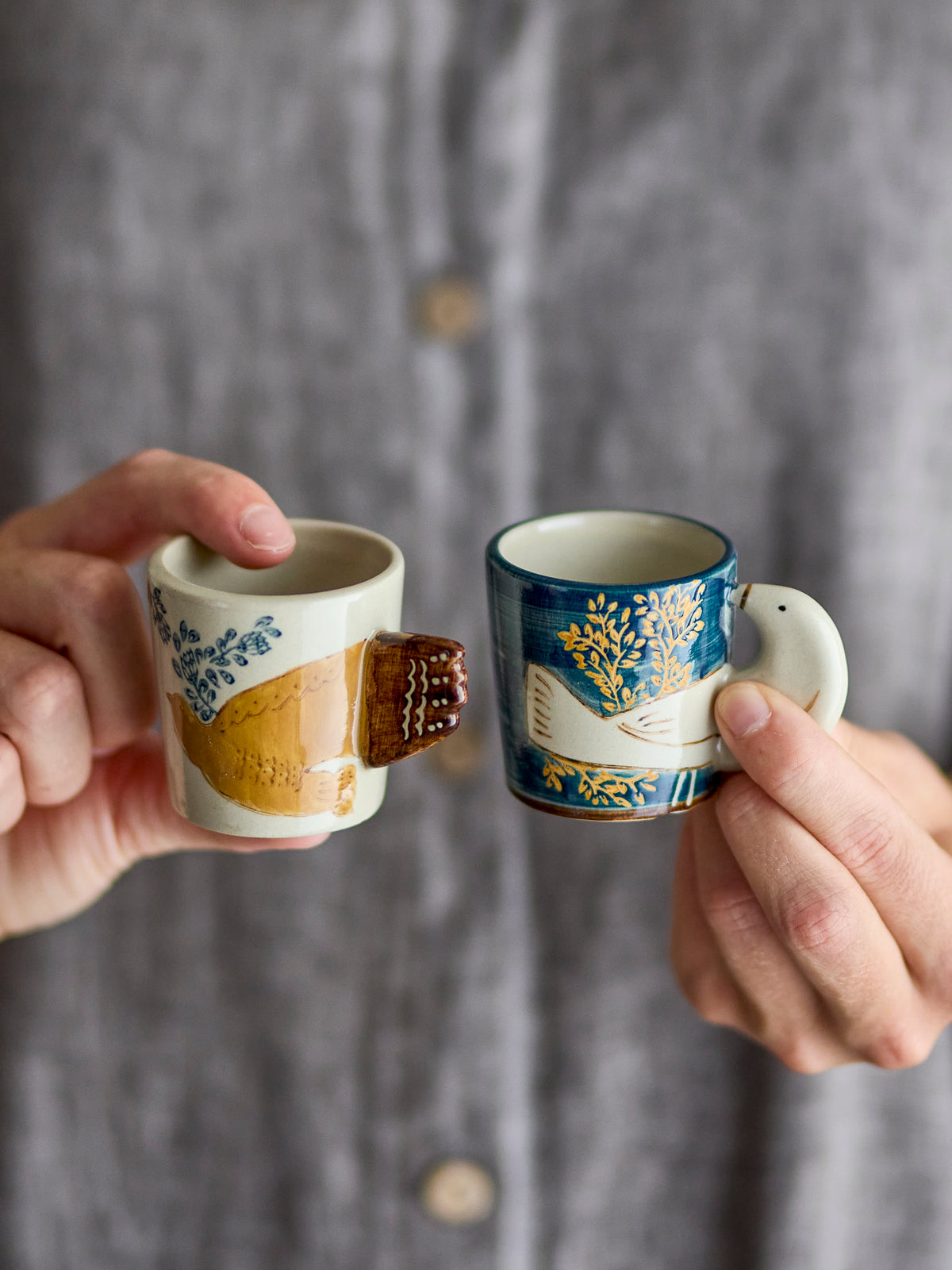 Two hands holding Hezha espresso cups, one cream with a golden bird motif and one navy with a golden floral pattern, against a grey knitted cardigan
