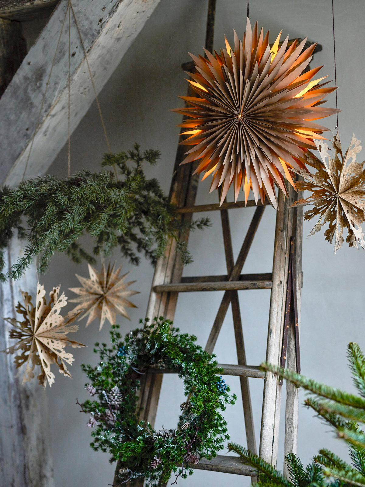 Cedar wreath hanging from the ceiling beside paper stars, a wooden ladder and a round green wreath in a rustic room