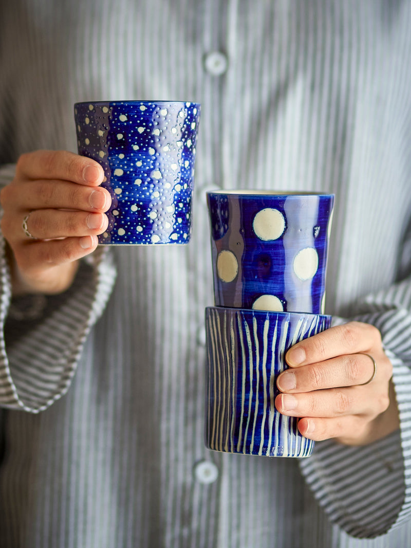 Minoa mugs in blue, Stripe, Dots, and Stars designs, shown in hand for scale
