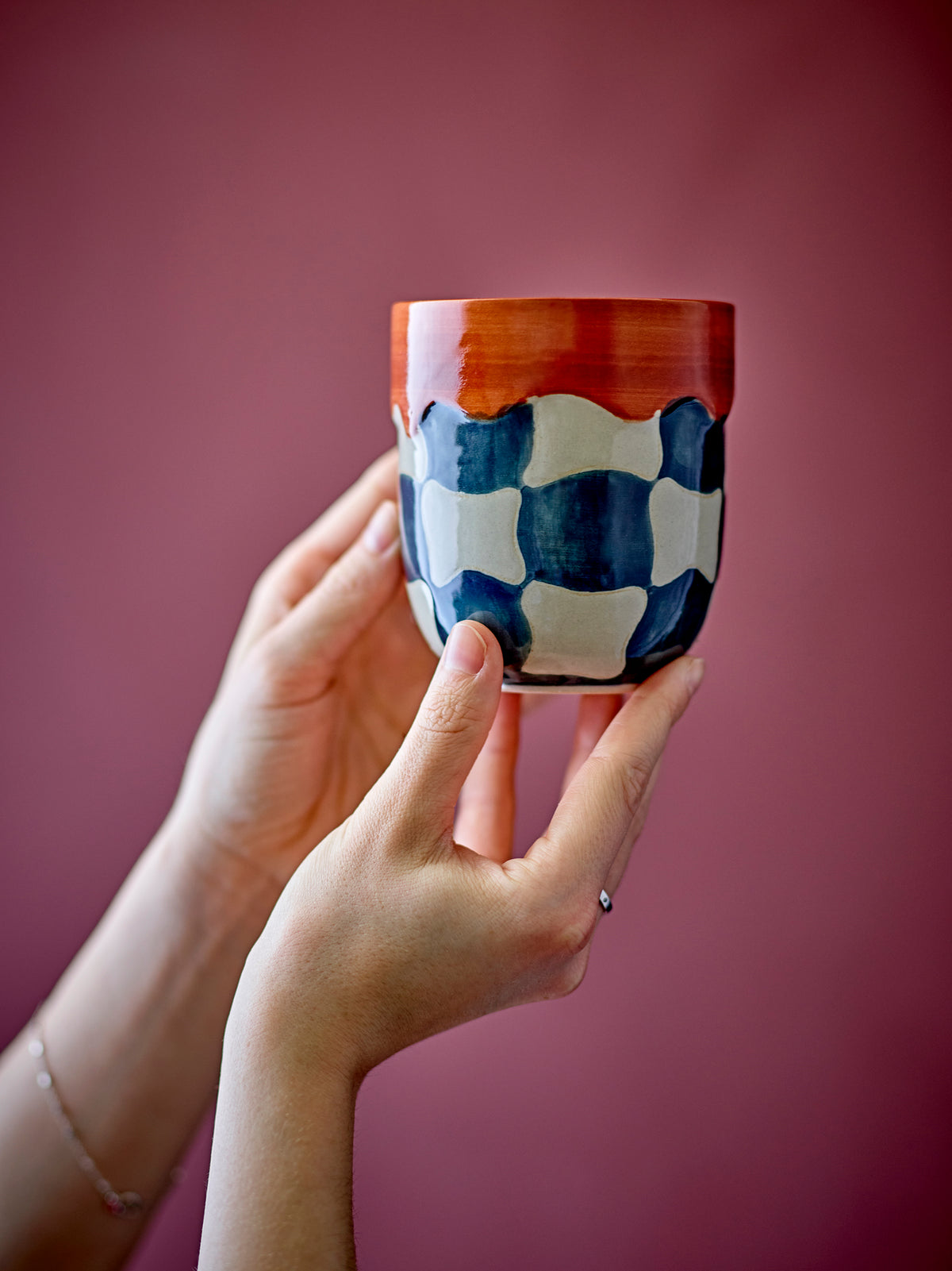 Check cup in blue and cream checkered stoneware with red rim, held in hand
