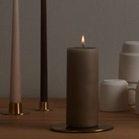 Large brown pillar candle alight on a gold candle plate with other, slimmer candles behind, and some cups stacked upside down